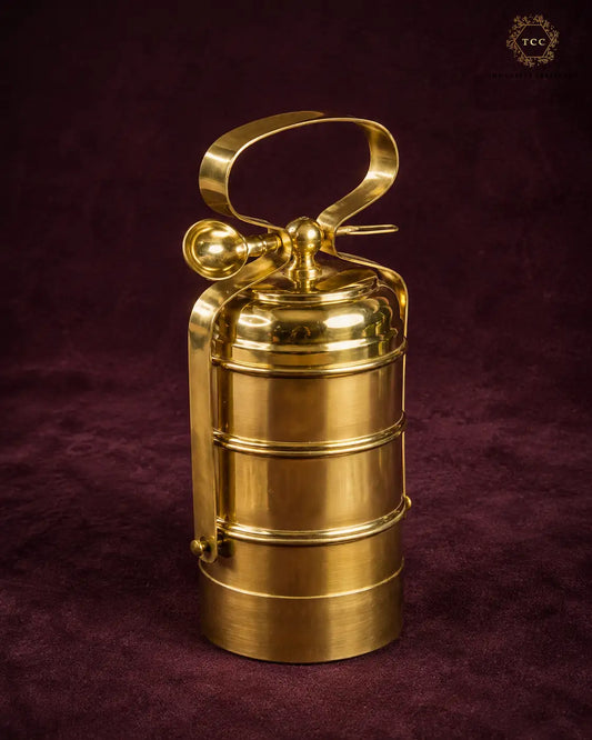 A polished brass fire extinguisher-shaped decorative object with a loop handle and cylindrical body, standing on a dark burgundy textured fabric background.