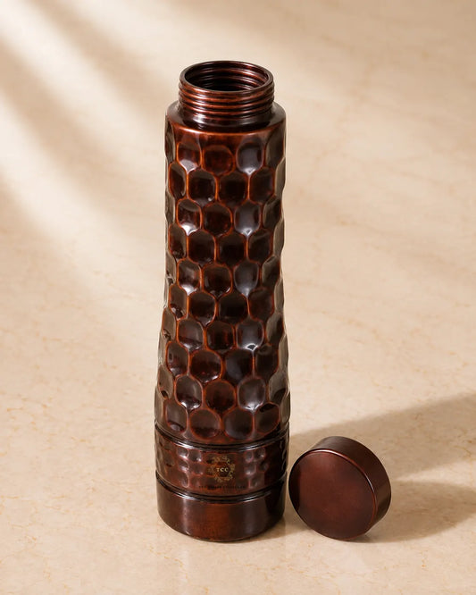 A hammered copper water bottle with a matching round cap placed beside it on a beige surface, shown in warm natural light with soft shadows.