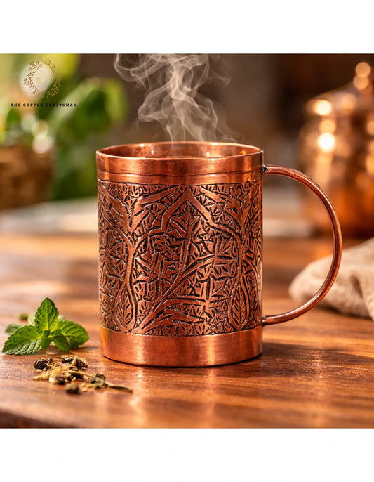 Copper mug with intricate patterns on a wooden surface, surrounded by mint leaves and tea leaves.