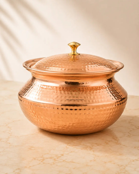 A hammered copper lidded serving pot with a small gold-toned knob on top, set on a light beige marble surface with soft shadow patterns on a cream background.