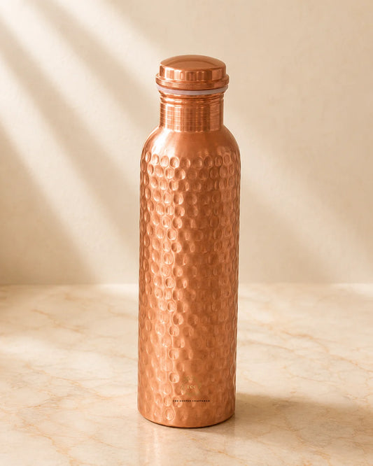 A hammered copper water bottle with a matching screw-on lid, standing on a light marble surface against a warm beige background with soft diagonal shadows.