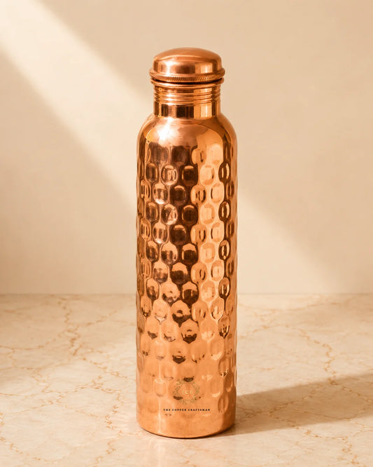 A copper water bottle with a hammered texture and a matching screw-on lid, standing on a light marble surface against a warm beige background.