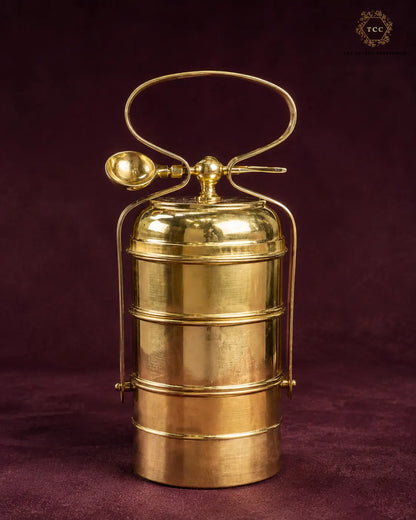 A polished brass 3 tiered tiffin box with a carrying handle and spoon, set against a dark burgundy background. A 'TTC' brand logo is visible in the top right corner.