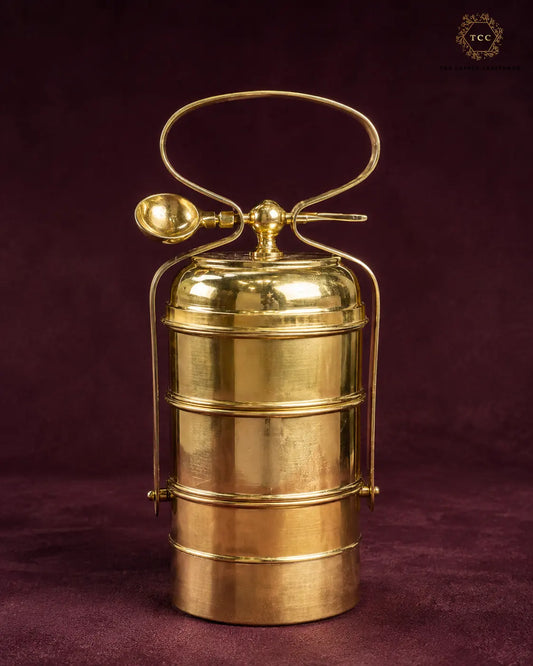 A polished brass 3 tiered tiffin box with a carrying handle and spoon, set against a dark burgundy background. A 'TTC' brand logo is visible in the top right corner.