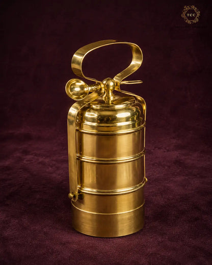A polished brass fire extinguisher-shaped decorative object with a loop handle and cylindrical body, standing on a dark burgundy textured fabric background.