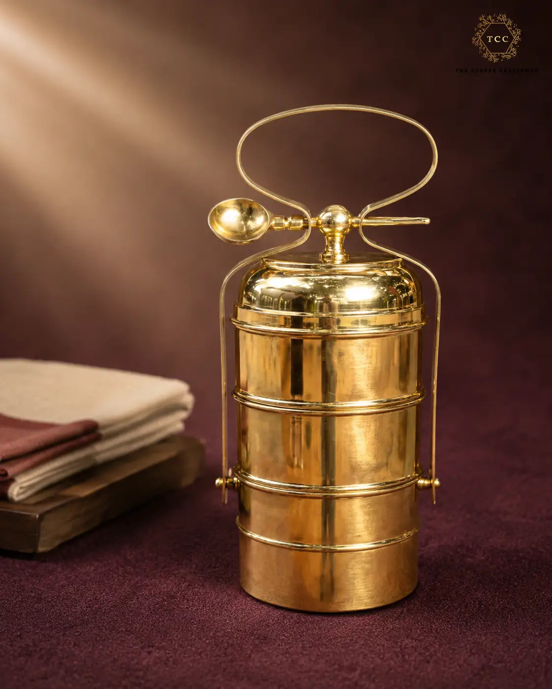 A polished brass tiered lunch box with a carrying handle and spoon, displayed on a dark purple textured surface next to stacked books, with a brand logo in the top right corner.
