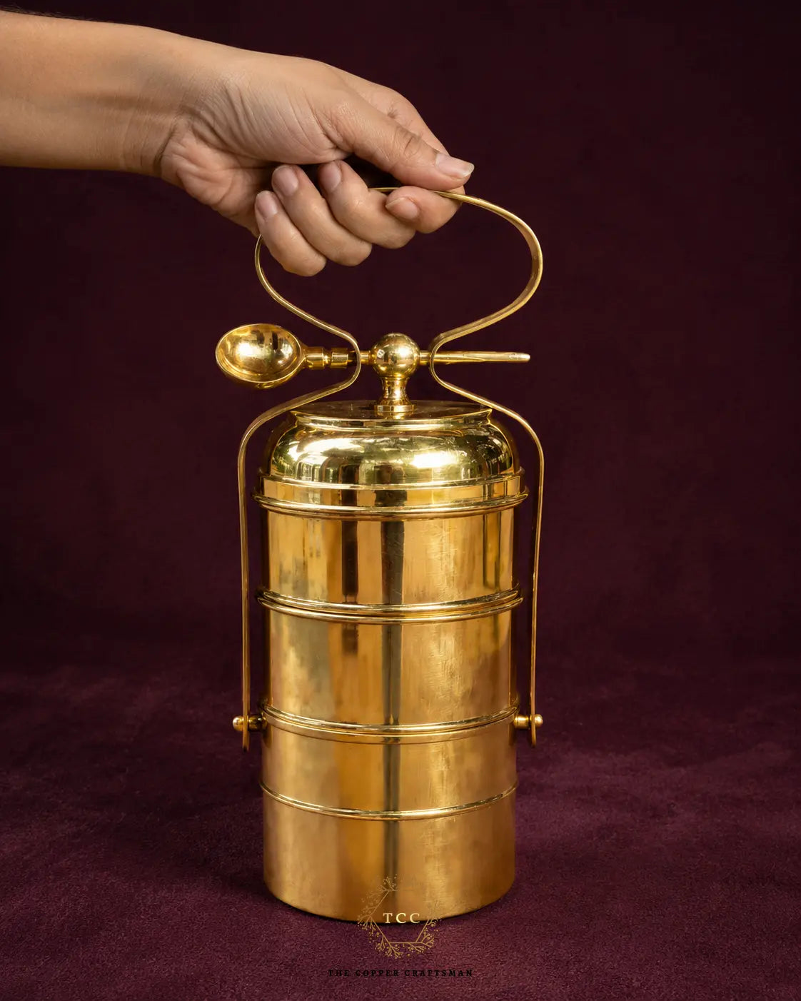 A hand holding the handle of a polished brass t3 iered tiffin box with a spoon lid, set against a dark burgundy background.