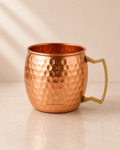 A hammered copper Moscow Mule mug with a polished brass handle, shown on a light beige marble surface with soft diagonal shadows.
