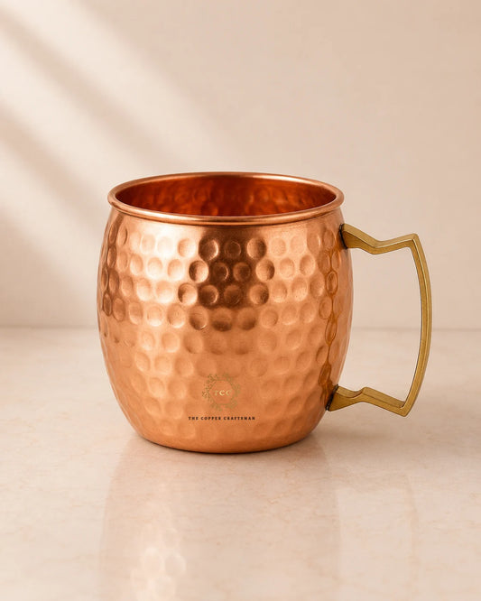 A hammered copper Moscow Mule mug with a polished brass handle, shown on a light beige marble surface with soft diagonal shadows.