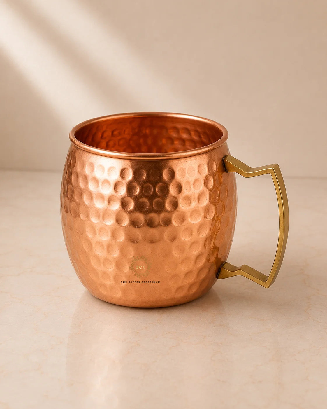 A hammered copper Moscow Mule mug with a polished brass handle, set on a light beige marble surface against a neutral beige background with soft diagonal shadows.