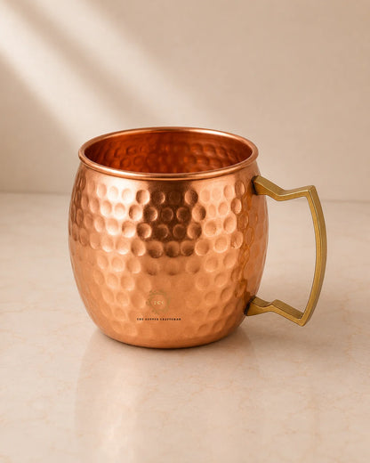 A hammered copper Moscow Mule mug with a polished brass handle, set on a light beige marble surface against a neutral beige background with soft diagonal shadows.