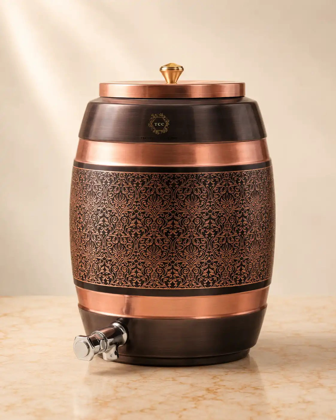 A Handcrafted Pure Copper Water Dispenser with an ornate embossed floral pattern on the front, a matching lid with a small gold finial, and a silver spigot, shown on a light stone surface against a warm beige background.