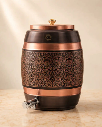 A Handcrafted Pure Copper Water Dispenser with an ornate embossed floral pattern on the front, a matching lid with a small gold finial, and a silver spigot, shown on a light stone surface against a warm beige background.