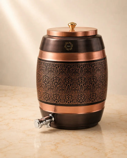 A Handcrafted Pure Copper Water Dispenserwith an ornate embossed pattern, a matching lid with a brass knob, and a metal spout, shown on a light beige marble surface against a warm beige background.