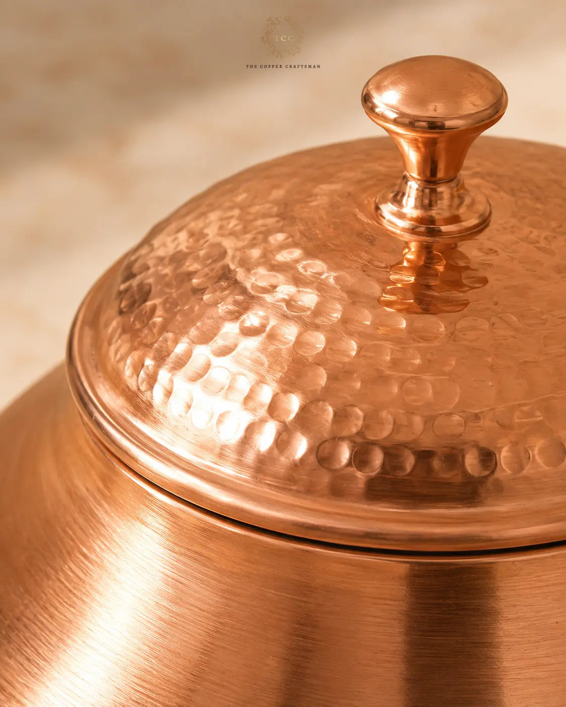 Close-up of a hammered copper lidded container with a rounded knob and brushed copper body, photographed in warm golden lighting against a softly blurred beige background.