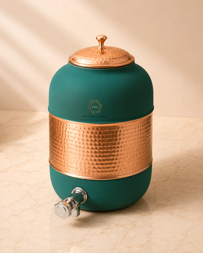 A round ceramic water dispenser in deep teal green with hammered copper-colored metal accents on the lid, midsection, and spout hardware, shown on a light beige marble surface with warm sunlight and shadows.