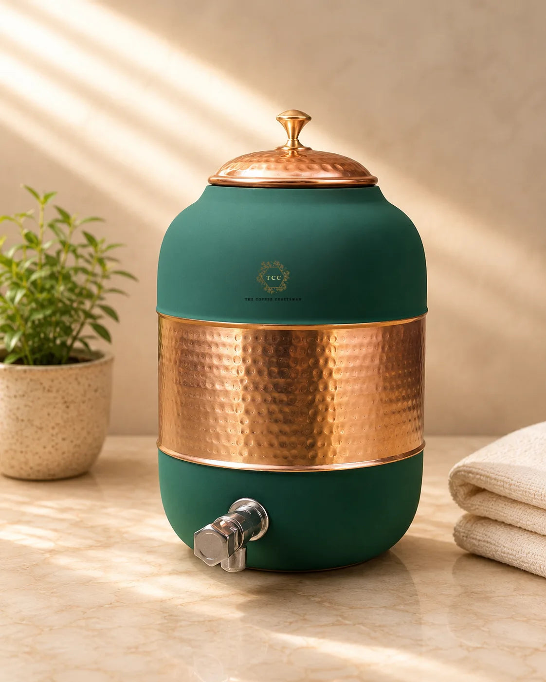 A round water dispenser with a matte dark green body, a hammered copper band around the middle, a matching copper lid, and a silver metal spigot. It sits on a beige marble surface with a small potted plant on the left and folded towels on the right, under warm diagonal sunlight.