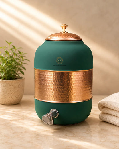 A round water dispenser with a matte dark green body, a hammered copper band around the middle, a matching copper lid, and a silver metal spigot. It sits on a beige marble surface with a small potted plant on the left and folded towels on the right, under warm diagonal sunlight.