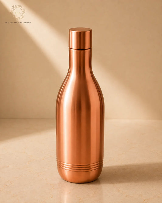 A copper-colored metal bottle with a matching lid, standing on a beige surface against a beige background with soft diagonal shadows. The brand name 'The Copper Aesthetics' is visible in the upper left corner.
