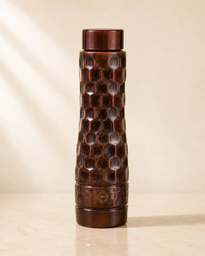 Antique Diamond Copper Water Bottle  with a honeycomb pattern and a matching lid, standing on a light beige marble surface against a warm beige background.