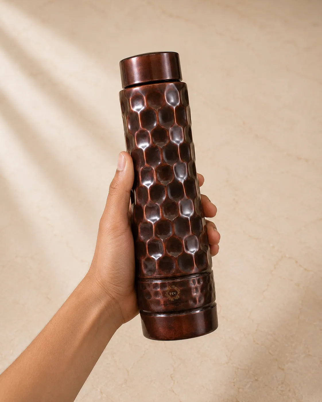A hand holding a Antique Diamond Copper Water Bottle with a honeycomb pattern, set against a warm beige textured background with soft leaf shadows.