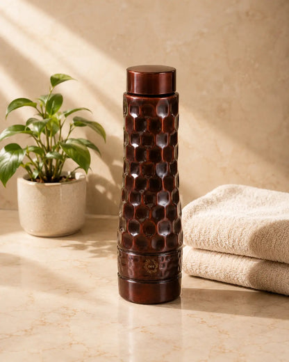 A dark brown Antique Diamond Copper Water Bottle with a honeycomb pattern, standing on a beige marble countertop next to a potted plant and folded towels.