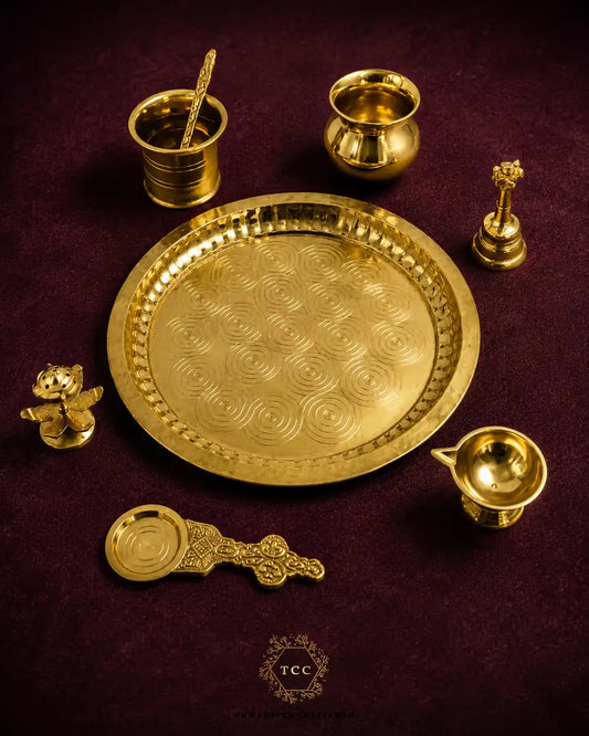 A set of brass Hindu ritual objects arranged on a dark purple textured background, including a large round plate, a small lamp, a spoon, a bowl, and decorative figurines. A 'TCC' logo is visible at the bottom.