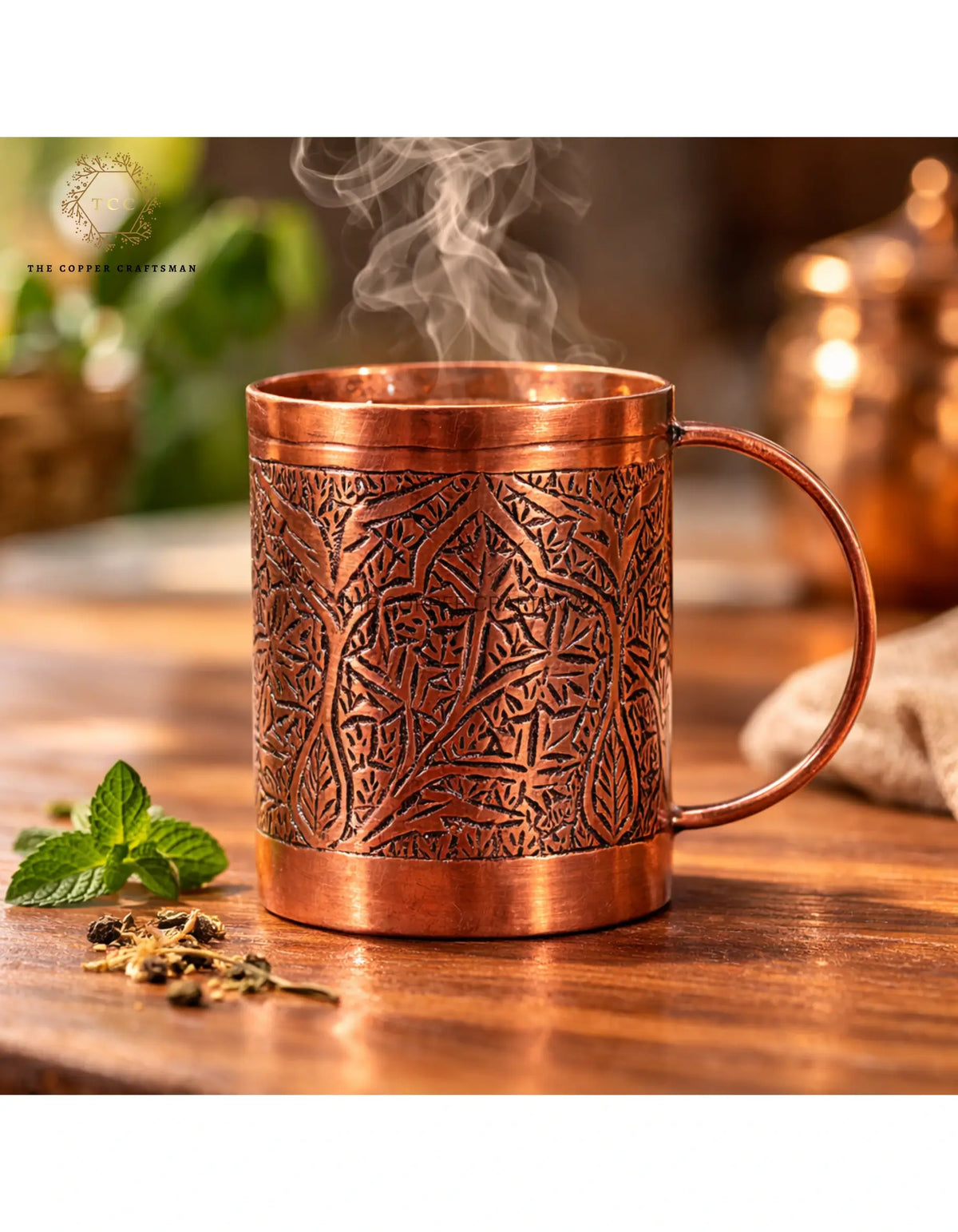 Copper mug with intricate patterns on a wooden surface, surrounded by mint leaves and tea leaves.