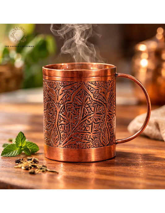Copper mug with intricate patterns on a wooden surface, surrounded by mint leaves and tea leaves.