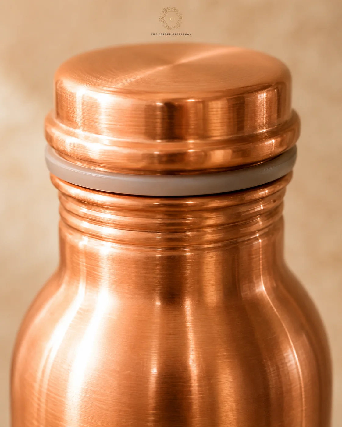 Close-up of a Classic 1 litre pure copper water bottle with a matching lid and a gray seal ring, shown against a warm beige background. A small logo at the top reads The Copper Collection.
