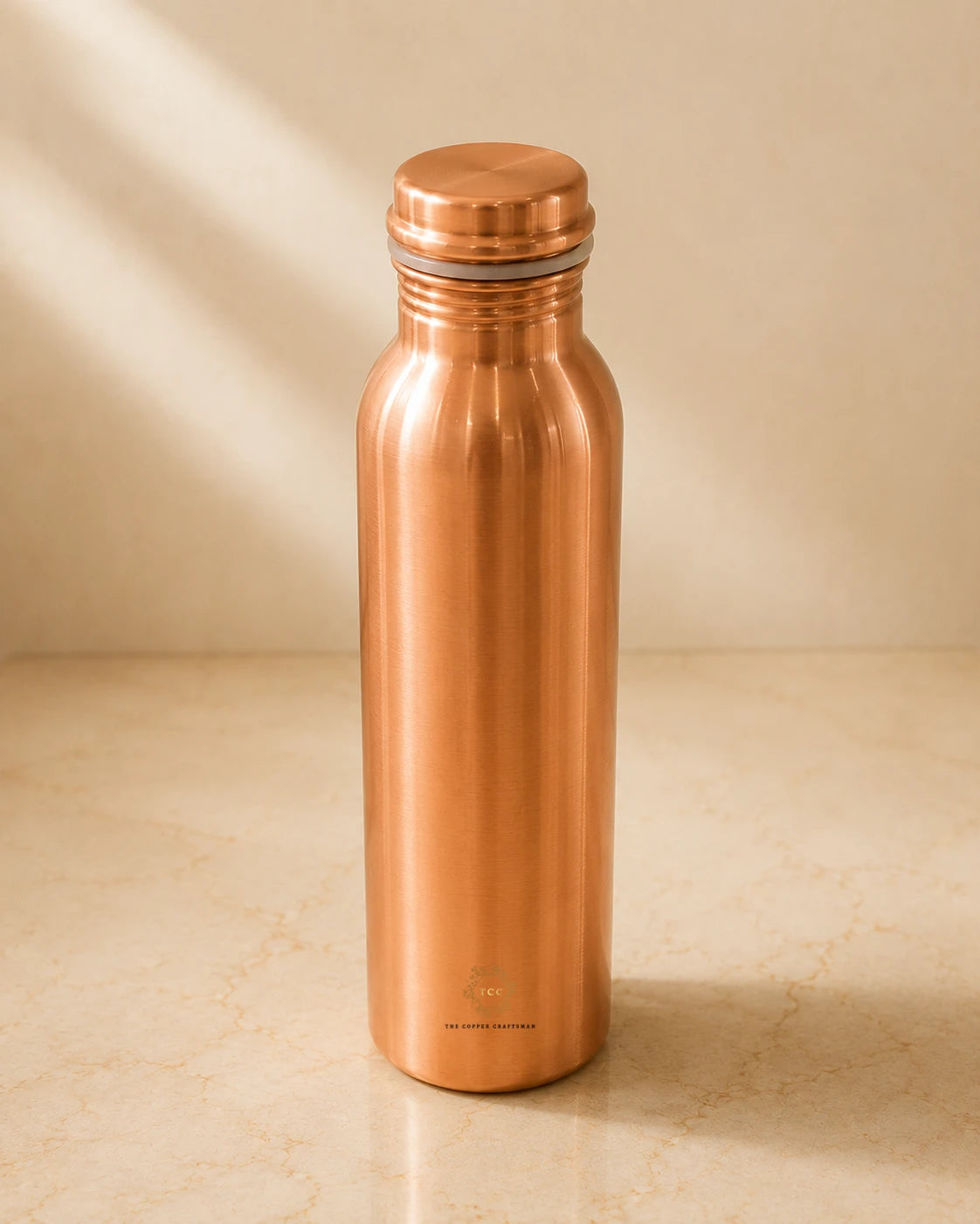 A Classic 1 litre pure copper water bottle with a matching screw-on lid, standing on a light beige marble surface against a matching beige background with soft diagonal shadows.