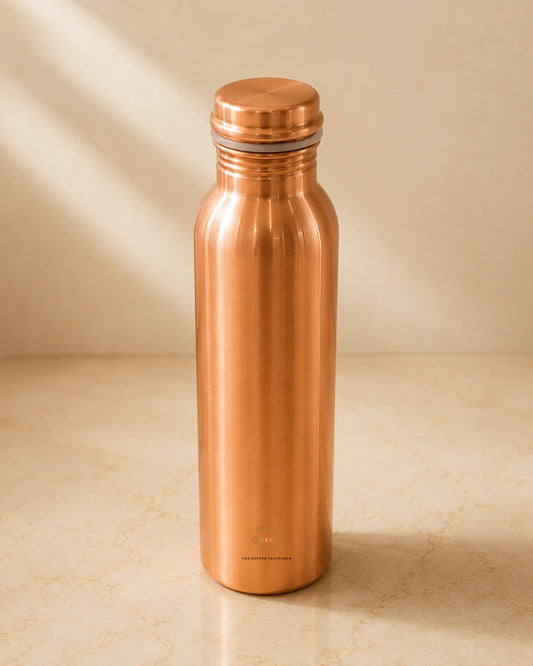 A Classic 1 litre pure copper water bottle with a matching screw-on lid, standing on a light beige marble surface against a matching beige background with soft diagonal shadows.