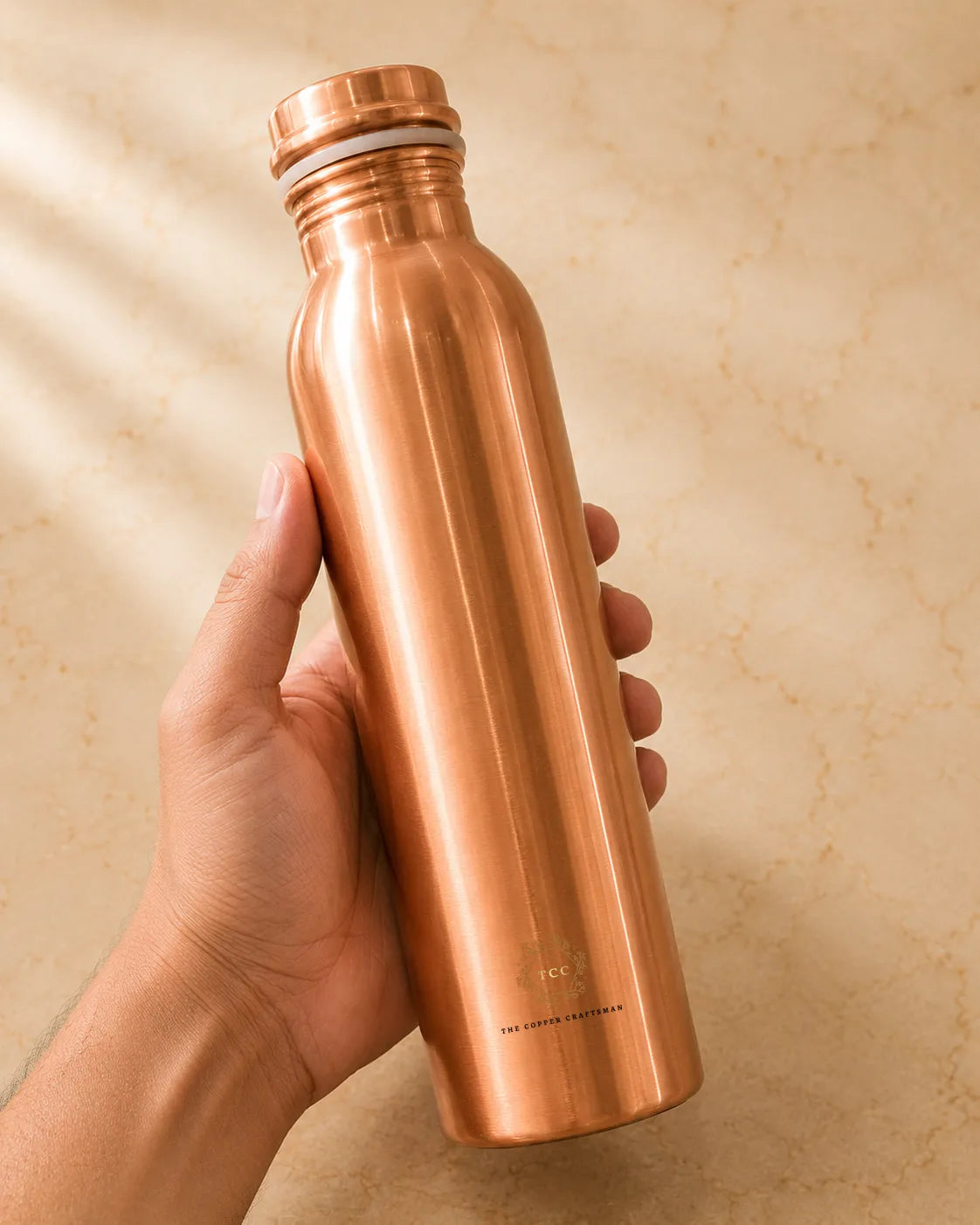 A hand holding a Classic 1 litre pure copper water bottle  against a warm beige stone-textured background.