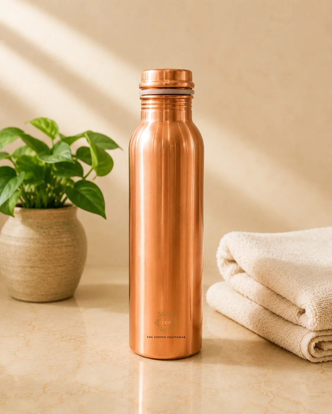 A Classic 1 litre pure copper water bottle with a screw cap stands on a beige marble surface, flanked by a potted green plant on the left and a folded cream towel on the right.