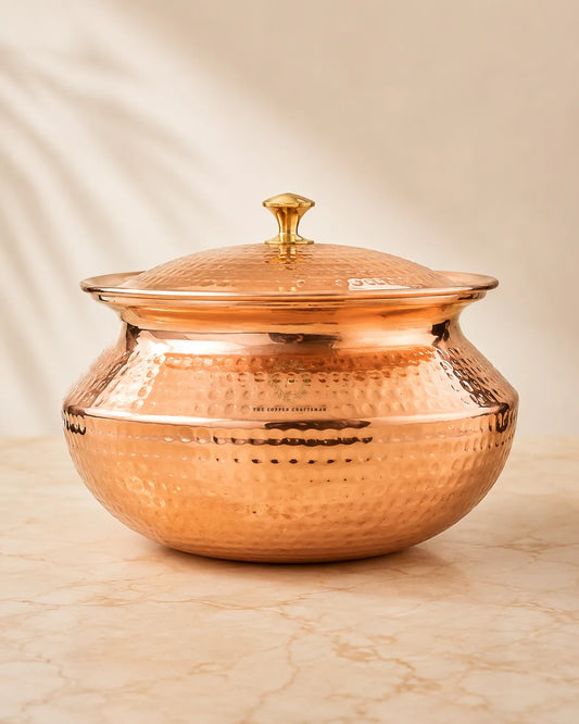 A hammered copper lidded pot with a gold-toned knob, sitting on a light beige marble surface against a soft beige backdrop.