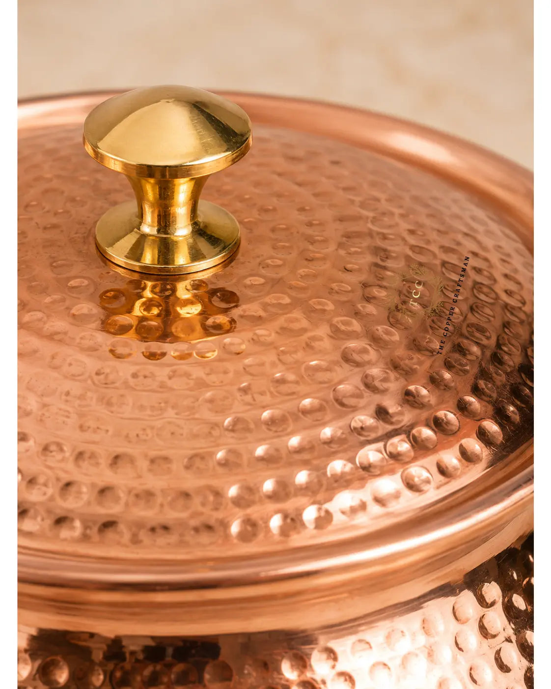 Close-up of a hammered copper pot lid with a polished brass knob, showing the textured metallic surface and warm copper tones against a softly blurred beige background.