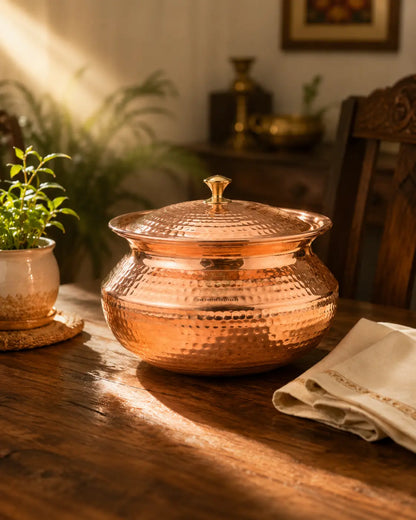 A hammered copper lidded pot with a small top knob, set on a rustic wooden table in warm sunlight with a potted plant and folded cloth nearby.