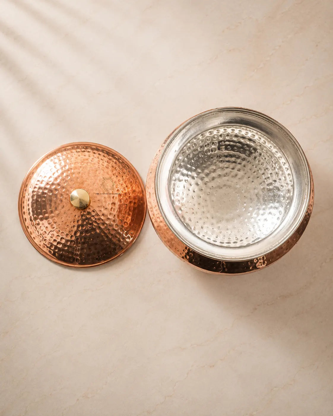 A hammered copper lidded container shown open, with the matching hammered copper lid placed beside it on a light beige surface.