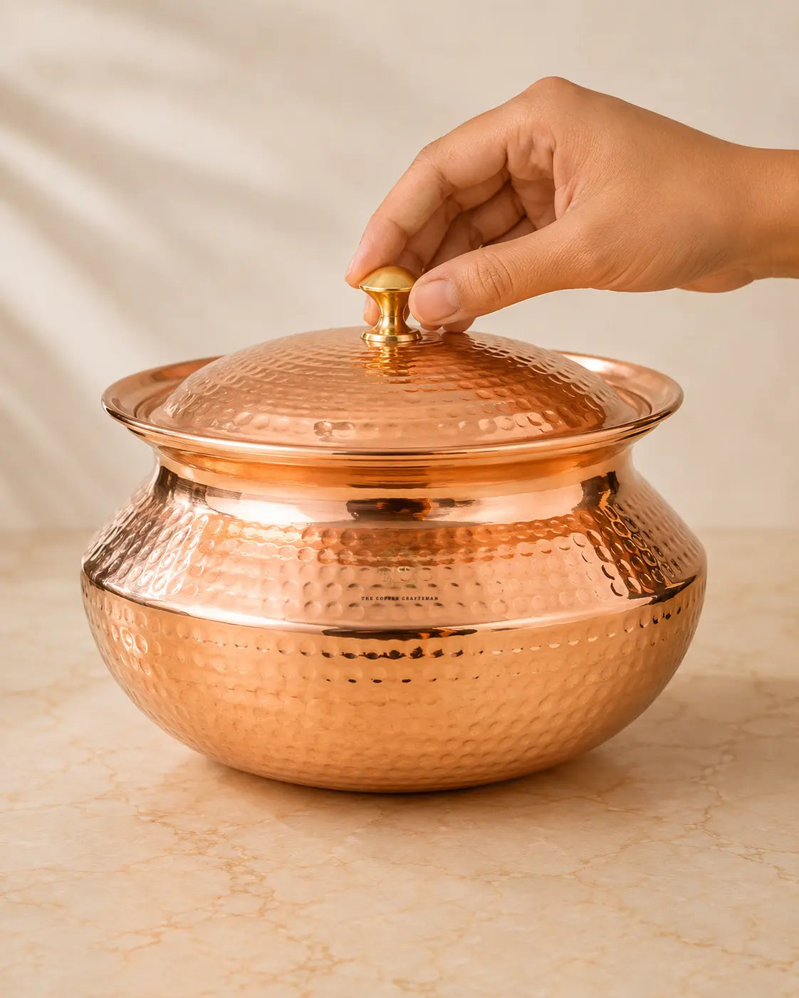 A hammered copper lidded pot with a small brass knob, shown on a light stone surface with a hand reaching to lift the lid.