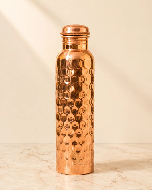 A hammered honeycomb copper water bottle with a matching lid, standing on a light marble surface against a warm beige background.