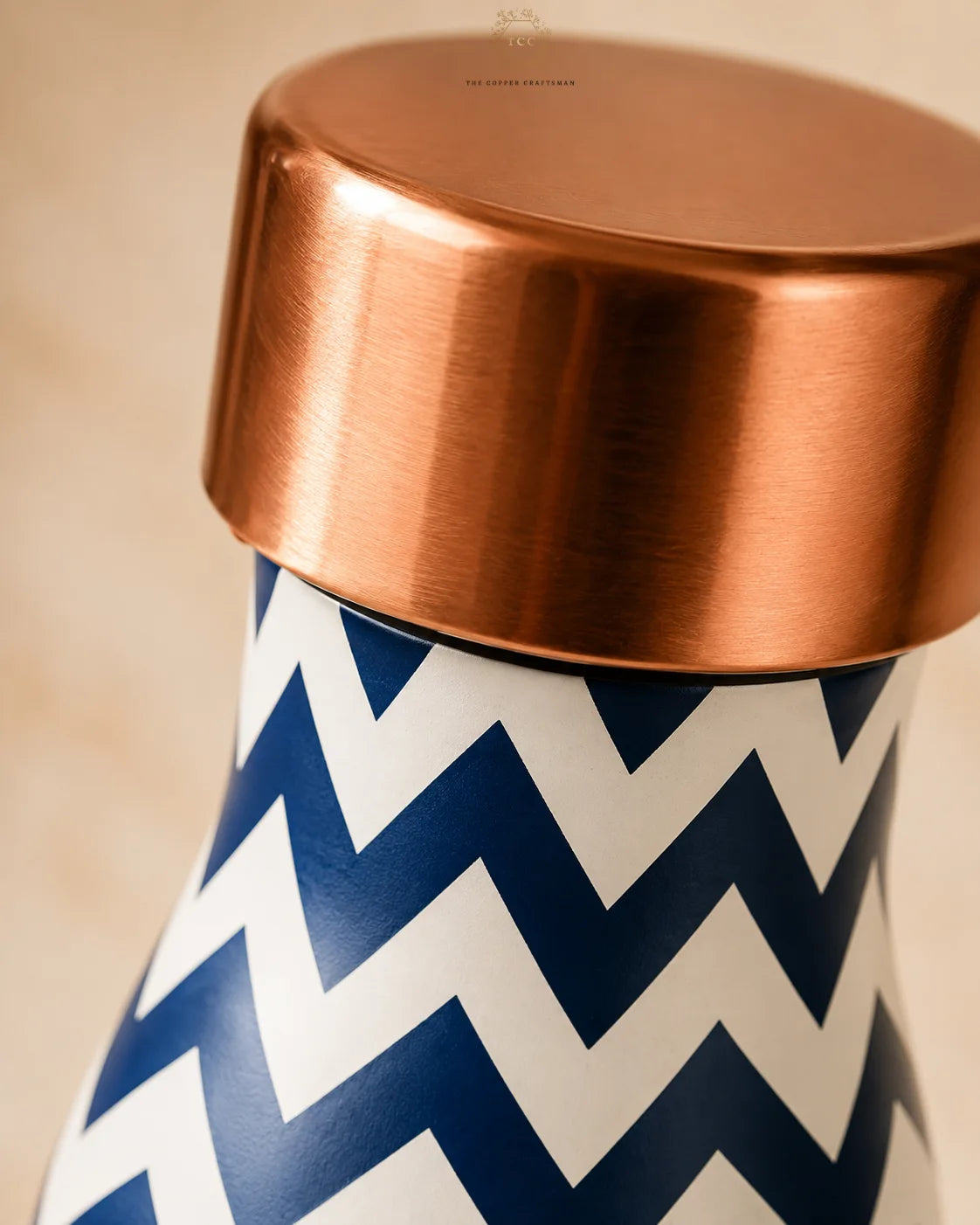 Close-up of a copper bottle or jar with a brushed copper lid and a navy blue and white chevron patterned body, photographed against a soft beige background.