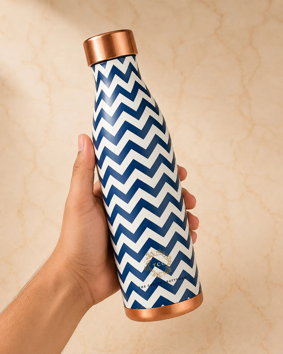 A hand holding a copper water bottle with a navy blue and white chevron pattern and metallic rose gold accents on the cap and base, set against a beige marble background.