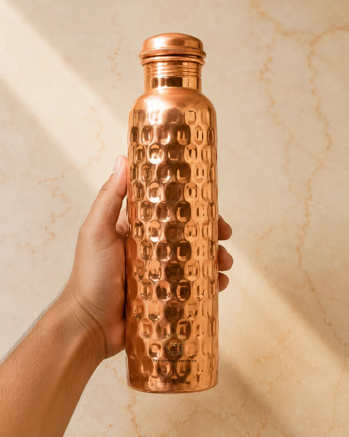 A hand holding a honycomb copper water bottle against a warm beige marble background.