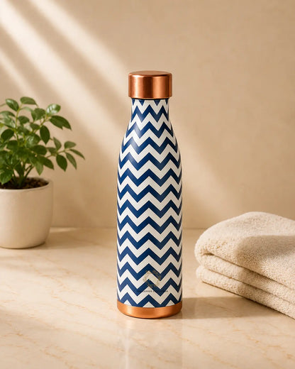 A tall copper water bottle with a navy and white chevron pattern and metallic copper-colored lid and base, standing on a beige marble surface next to a small potted plant and folded towels.