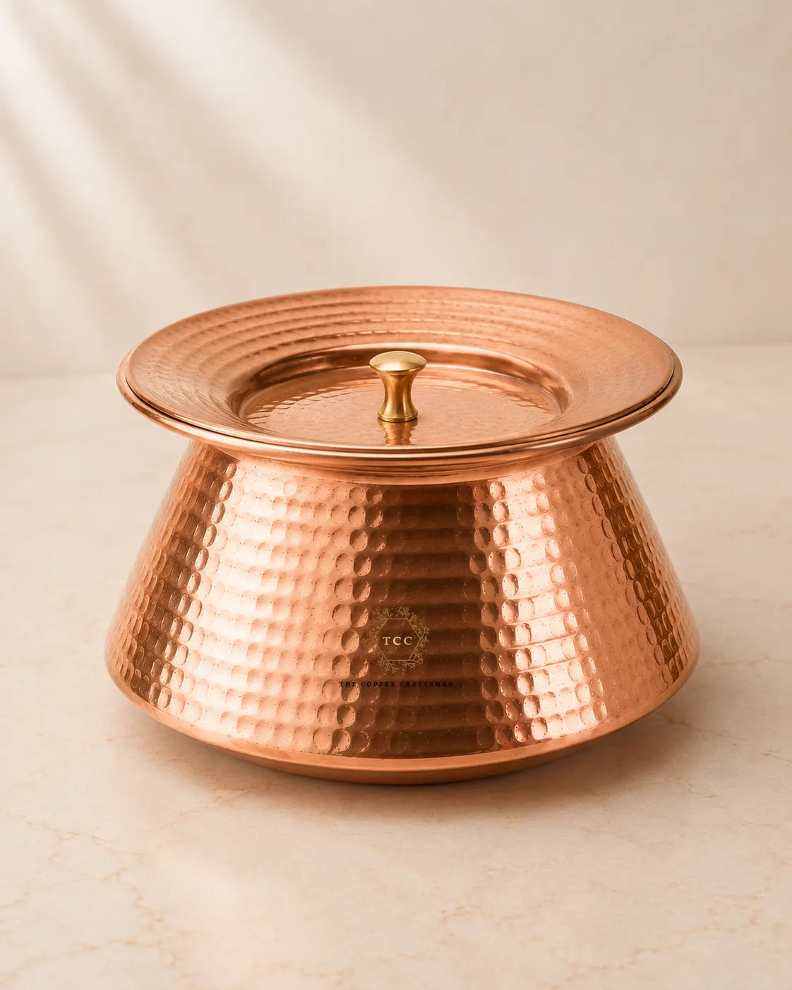 A hammered copper lidded handi with a matching lid and small central knob, photographed on a light beige marble-like surface against a soft neutral background.