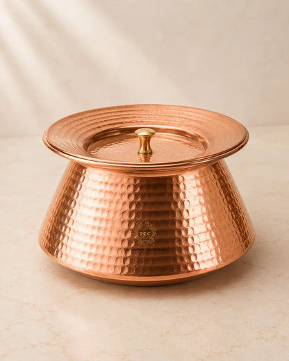 A hammered copper lidded handi with a matching lid and small central knob, photographed on a light beige marble-like surface against a soft neutral background.
