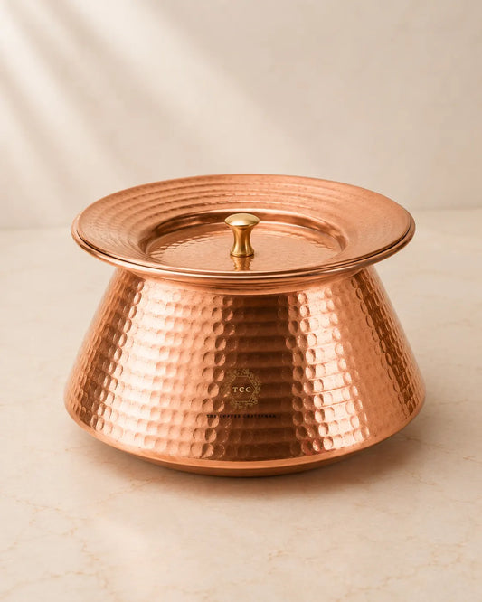 A hammered copper lidded handi with a matching lid and small central knob, photographed on a light beige marble-like surface against a soft neutral background.