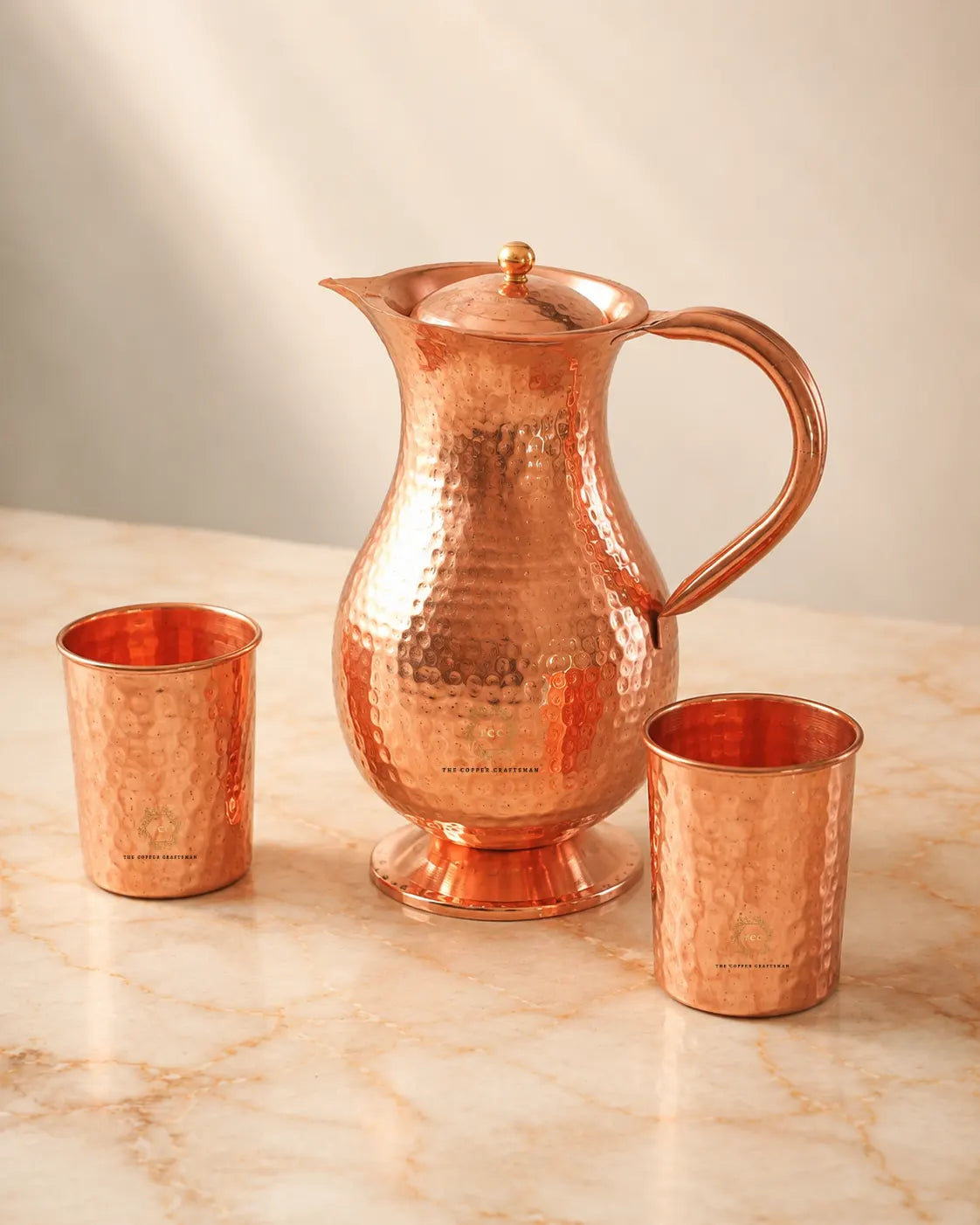 A hammered copper pitcher with a lid and two matching copper tumblers, arranged on a light marble surface against a soft beige background.