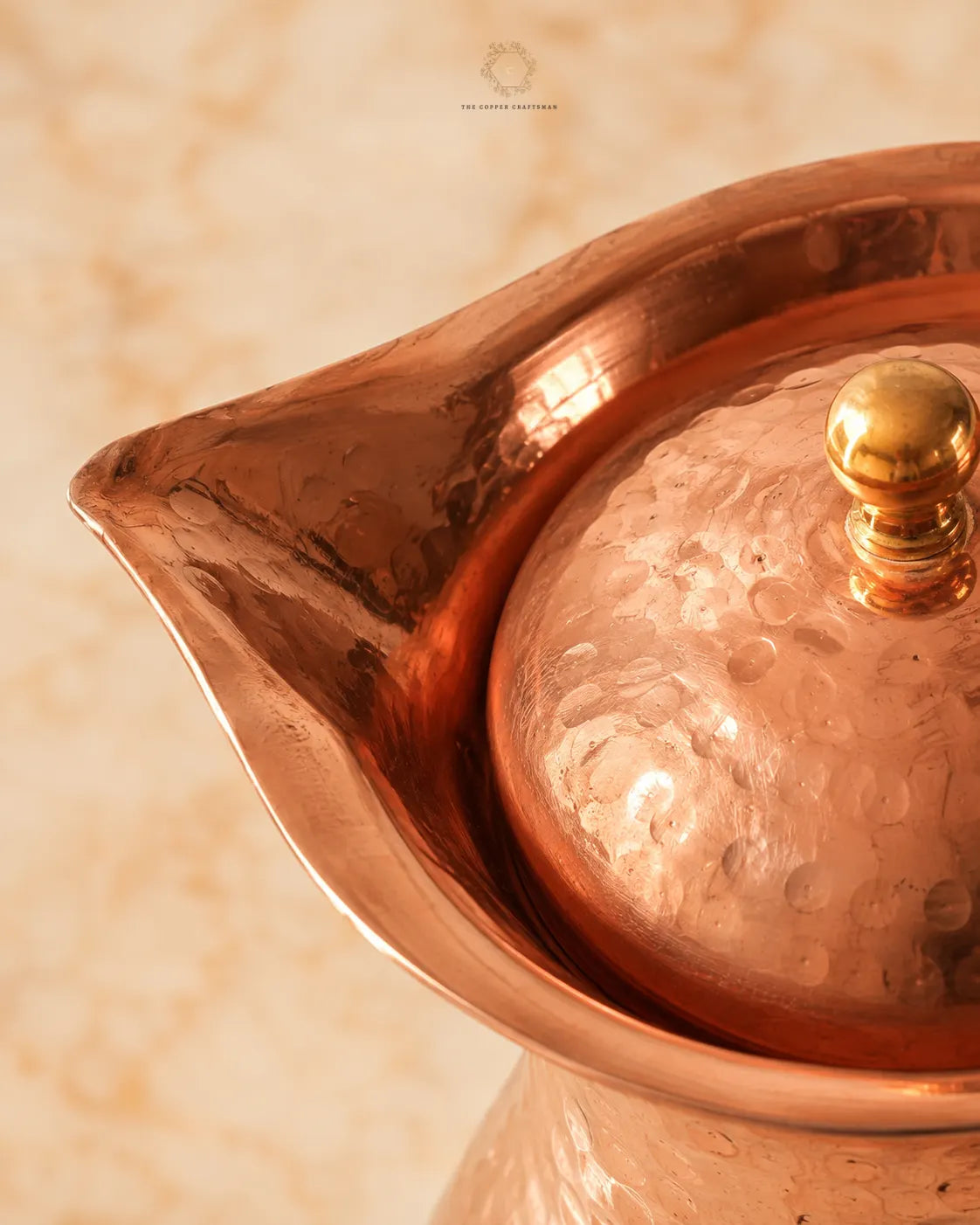 Close-up of a hammered copper teapot or pitcher with a matching copper lid and brass-toned knob, set against a warm beige marble-like background.
