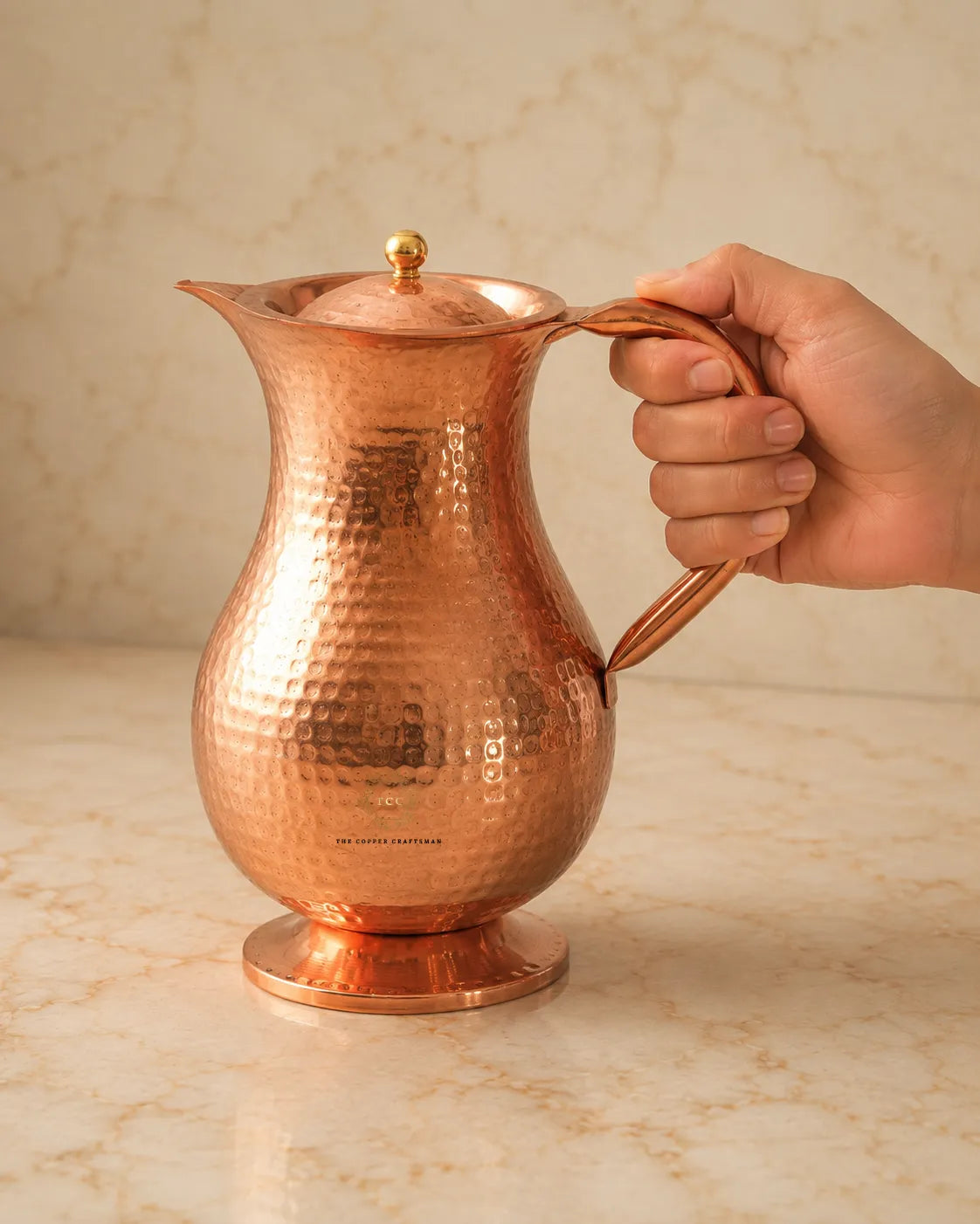 A hammered copper water pitcher with a matching lid and a curved handle, shown with a hand holding the handle on a beige marble surface and background.